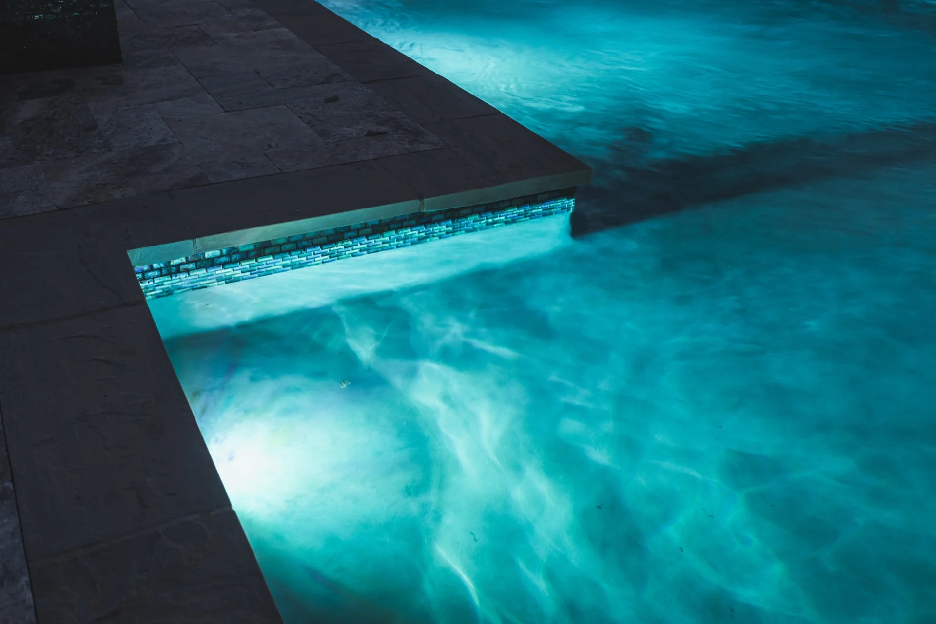 LED-lit pool edge with glass tile band and travertine coping at night — Tarkington Residence